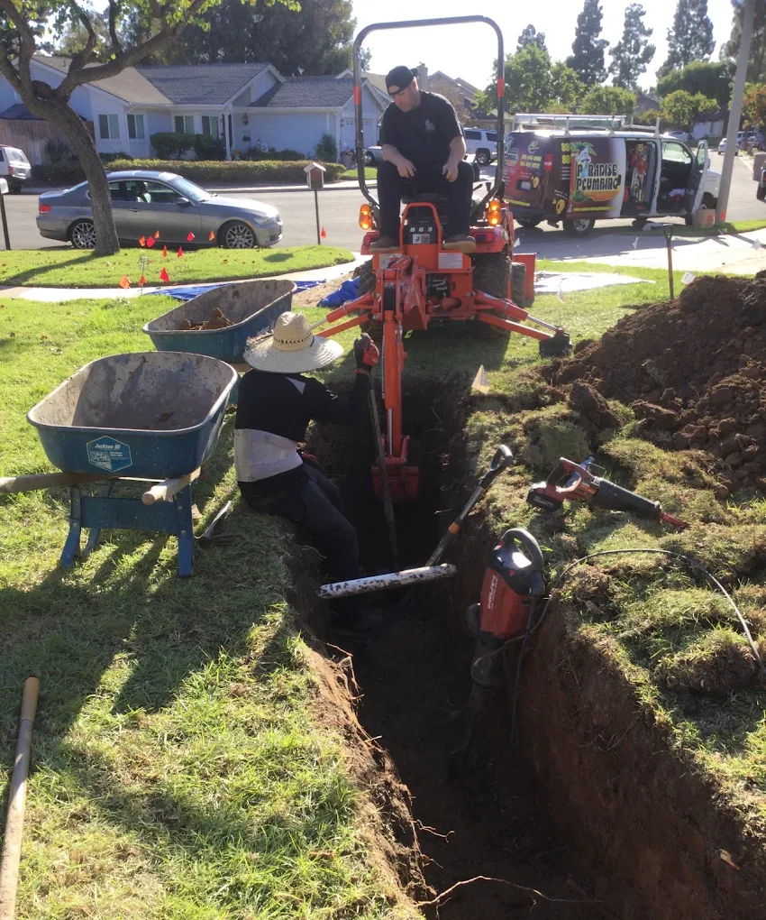 AAA Paradise Plumbing crew running a service line trench in Ventura, CA &mdash; one technician operating a mini excavator while another works in the open trench, branded Paradise Plumbing van parked behind